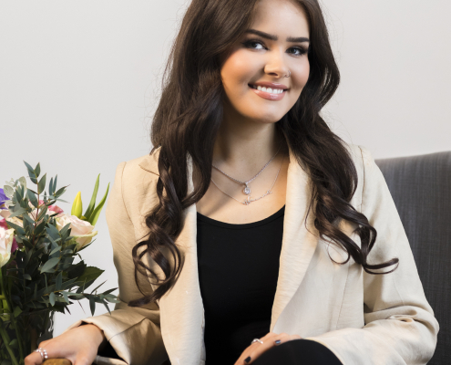 A woman with long brown hair is wearing a black top and trousers with a cream blazer. She is sat on a chair in front of a grey wall beside flowers.